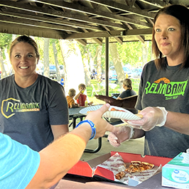 Two Reliabank employees smile while one hands a plate with pizza to an individual at a free community event hosted by Reliabank.