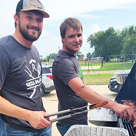 Two Reliabank employees smile standing at a grill, one holding a tongs with a hot dog, at an event serving Hamlin County community teachers.