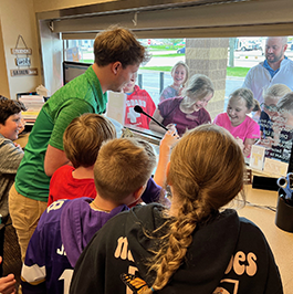 Fourth grade students surround a drive-up teller as he demonstrates how the drive-up banking system works by using the microphone to talk to another Reliabank employee and group of students outside the window.